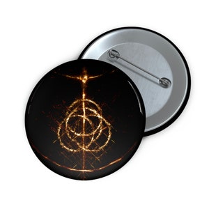 Könnte beinhalten: Zwei runde, schwarze Knöpfe mit metallischer Stiftunterseite. Der vordere Knopf zeigt ein leuchtendes goldenes Design auf schwarzem Hintergrund. Das Design ist ein kompliziertes, abstraktes Symbol mit einer zentralen vertikalen Linie und kreisförmigen Elementen.