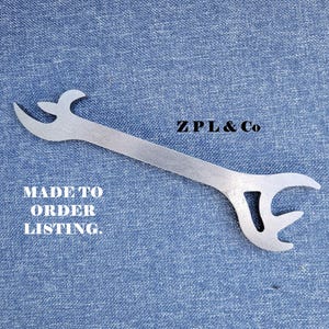 Może przedstawiać: Srebrny metalowy klucz z zakrzywionymi końcami i centralnym otworem, spoczywający na niebieskiej powierzchni z dżinsu. Widoczny tekst "ZPL & Co" i "MADE TO ORDER LISTING".