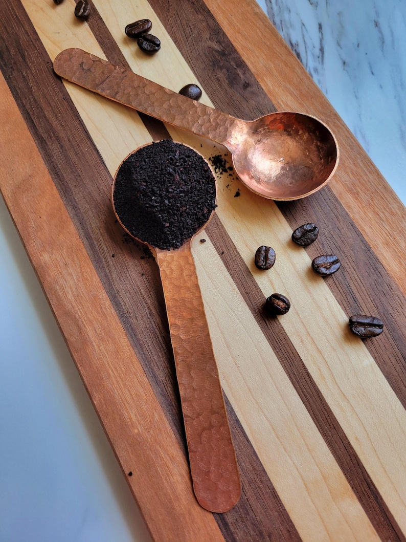 May include: Two copper coffee scoops, one filled with dark coffee grounds, rest on a striped wooden cutting board. Coffee beans are scattered around the scoops. The scoops have a hammered texture and a warm, reddish-brown color.