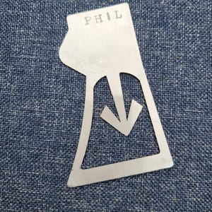 May include: A metal template with the word "PHIL" engraved on it. The template is shaped like a downward pointing arrow.