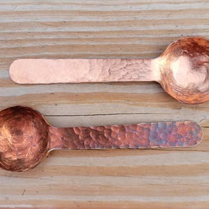 May include: Two copper spoons with hammered textures rest on a wooden surface. The spoons have rounded bowls and elongated handles. The copper has a warm, reddish-brown hue, with variations in color due to the hammering process.