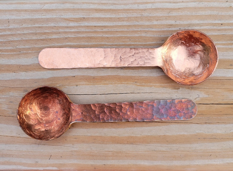 May include: Two copper spoons with hammered textures rest on a wooden surface. The spoons have rounded bowls and elongated handles. The copper has a warm, reddish-brown hue, with variations in color due to the hammering process.