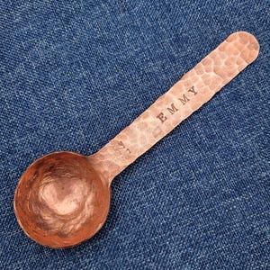 May include: A copper measuring spoon with a hammered texture. The handle is stamped with the word "EMMY". The spoon has a rounded bowl and is resting on a blue denim surface. The spoon is designed for measuring dry ingredients.