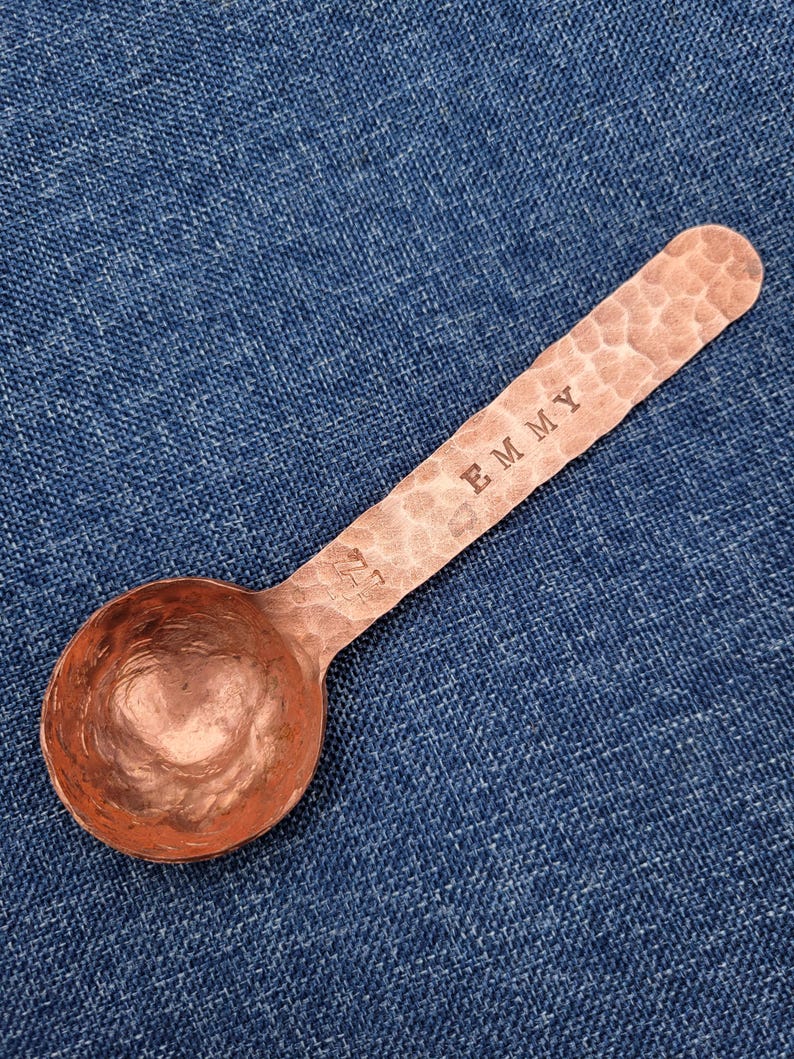 May include: A copper measuring spoon with a hammered texture. The handle is stamped with the word "EMMY". The spoon has a rounded bowl and is resting on a blue denim surface. The spoon is designed for measuring dry ingredients.