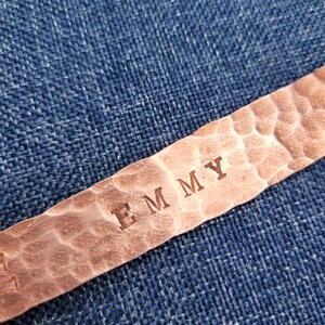 May include: Close-up of a copper bracelet with the name "EMMY" stamped into the metal. The bracelet has a hammered texture and is set against a blue denim background.