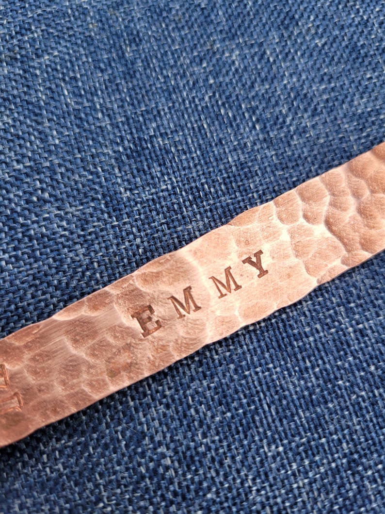 May include: Close-up of a copper bracelet with the name "EMMY" stamped into the metal. The bracelet has a hammered texture and is set against a blue denim background.