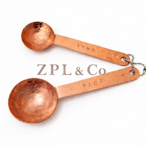 May include: Two copper measuring spoons with the word "TINA" stamped on the handle. The spoons are connected by a small chain. The text "ZPL & Co" is in the center of the image.