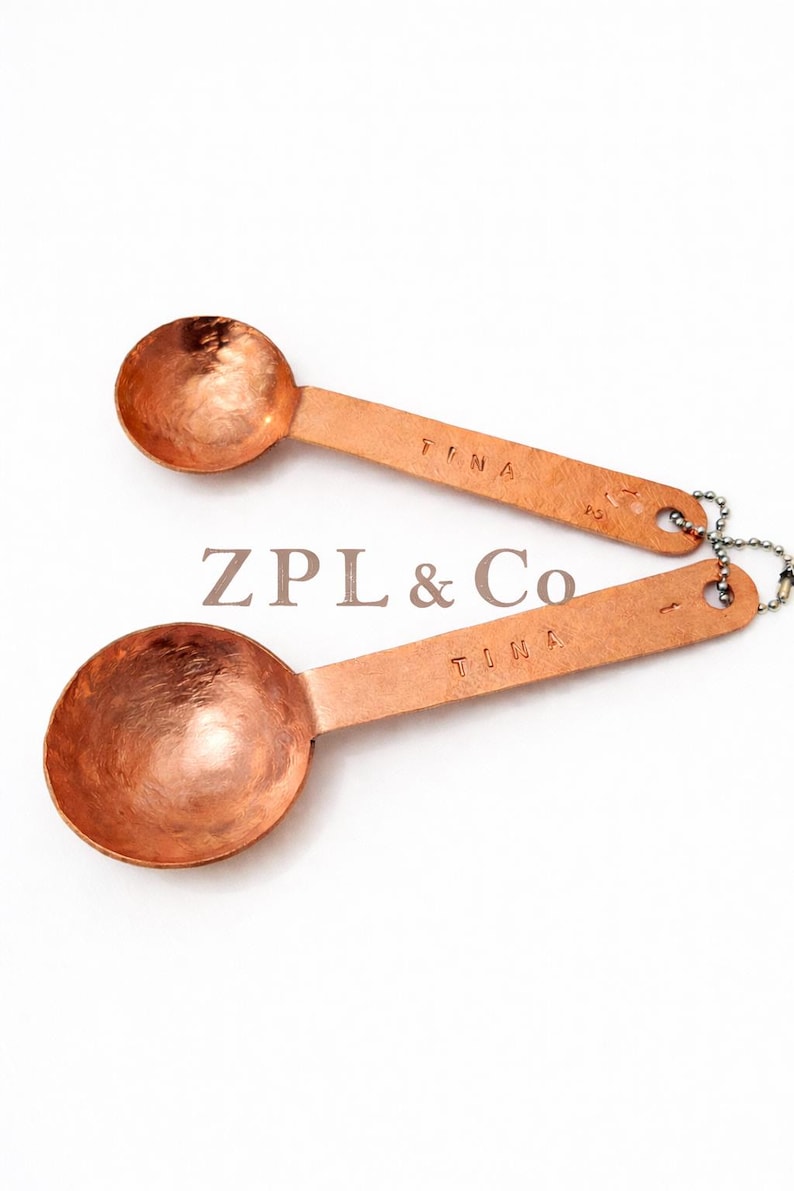 May include: Two copper measuring spoons with the word "TINA" stamped on the handle. The spoons are connected by a small chain. The text "ZPL & Co" is in the center of the image.