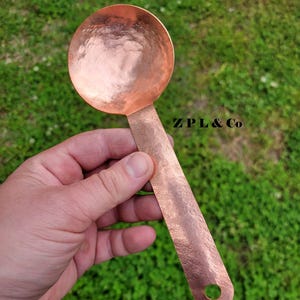 May include: A copper-colored ladle with a hammered texture. The ladle has a round bowl and a long, flat handle with a hole at the end. The text "ZPL & Co" is visible on the handle.