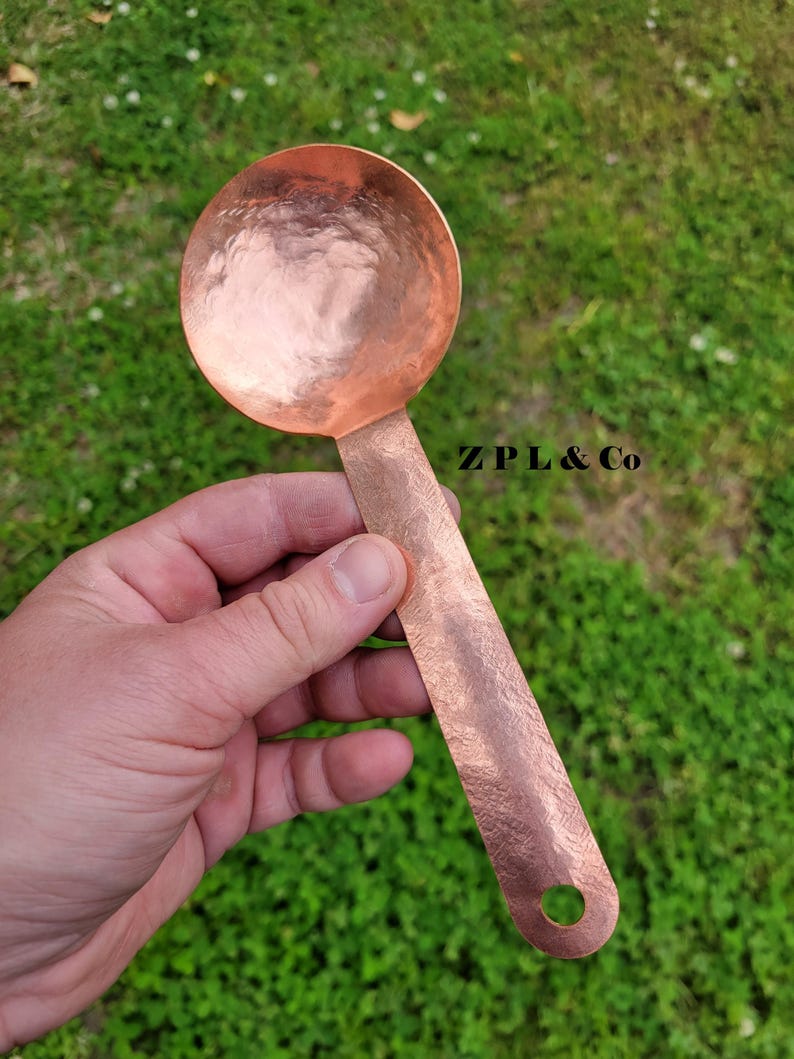 May include: A copper-colored ladle with a hammered texture. The ladle has a round bowl and a long, flat handle with a hole at the end. The text "ZPL & Co" is visible on the handle.