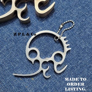 Puede incluir: Un llavero de metal plateado con un diseño estilizado único, con formas circulares y curvas. El llavero está unido a una pequeña cadena de bolas. Los textos "ZPL & Co" y "MADE TO ORDER LISTING" son visibles.