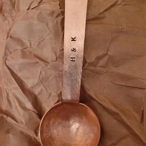 May include: A copper measuring spoon with a hammered texture. The spoon has a round bowl and a long handle with a hole at the end. The handle is stamped with the letters "H & K".