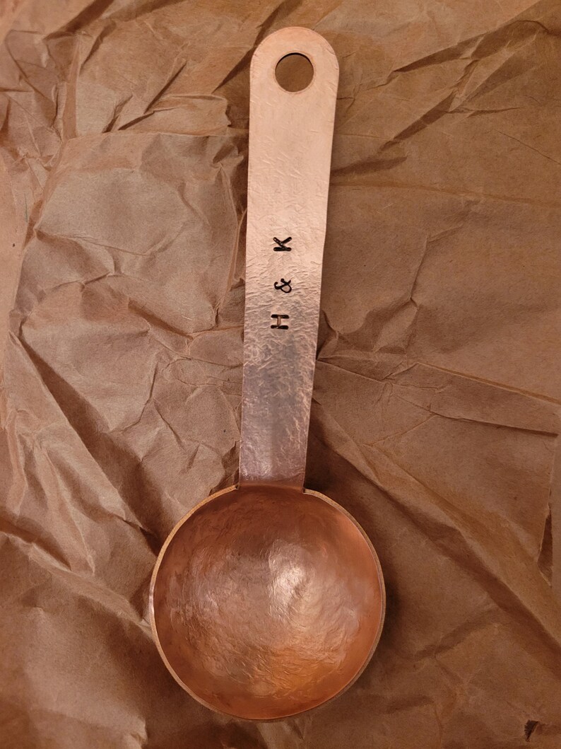 May include: A copper measuring spoon with a hammered texture. The spoon has a round bowl and a long handle with a hole at the end. The handle is stamped with the letters "H & K".
