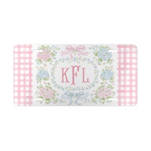 Op de afbeelding: Rechthoekige nummerplaat met een witte achtergrond en roze ruitjesranden. Het midden heeft een bloemenkrans met roze en blauwe hortensia's, een roze strik en de letters "KFL" in roze. Onderaan zit een blauwe strik.