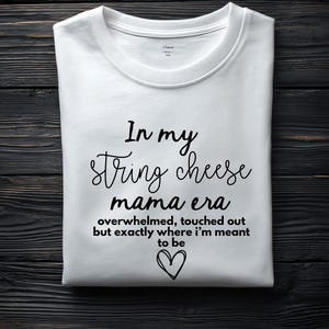 May include: White t-shirt with the text "In my string cheese mama era overwhelmed, touched out but exactly where I'm meant to be" in black script. A heart graphic is below the text. The shirt is folded.