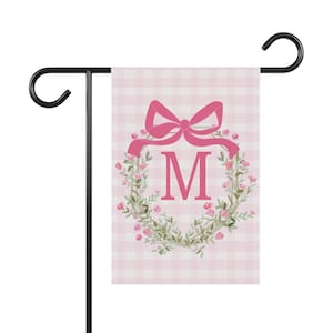 May include: A pink and white gingham garden flag with a pink floral wreath and a pink bow. The letter "M" is in the centre of the wreath.