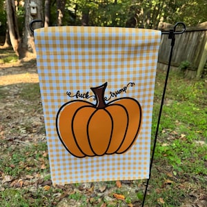 May include: A garden flag with a yellow and white gingham pattern. The flag features a large orange pumpkin graphic with the words "fuck trump" in black script. The flag is displayed outdoors on a black metal stand.