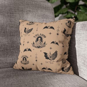 May include: A tan square pillow with a repeating black pattern of bats, mountains, stars, and text. The text includes "VELARIS CITY OF STARLIGHT" and "High lady of the night". The pillow is resting on a gray couch.