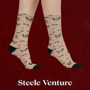 May include: A pair of beige socks with a repeating pattern of bats, stars, and other celestial elements. The socks have black heels and toes. The text "Steele Venture" is at the bottom of the image.