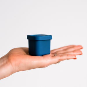 Puede incluir: Un pequeño recipiente azul cuadrado con una tapa a juego. El recipiente se sostiene en la palma de una mano sobre un fondo blanco. El recipiente parece estar hecho de un material blando.