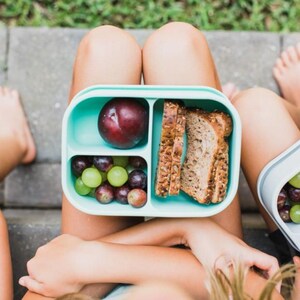 May include: A light teal lunchbox with three compartments, filled with a plum, grapes, and sliced bread. The lunchbox is open, revealing the contents. The image is taken from a top-down perspective.