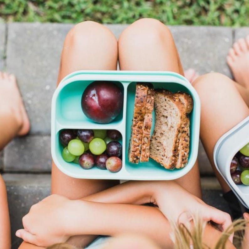 May include: A light teal lunchbox with three compartments, filled with a plum, grapes, and sliced bread. The lunchbox is open, revealing the contents. The image is taken from a top-down perspective.