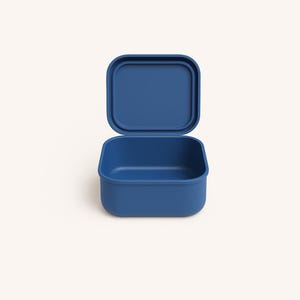 Puede incluir: Una fiambrera azul, de forma cuadrada, con tapa a juego, abierta para revelar el interior. La fiambrera está hecha de un material mate y liso. La tapa está ligeramente entreabierta, mostrando el interior del recipiente.