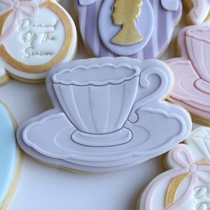 Puede incluir: Surtido de galletas de azúcar decoradas en colores pastel. Los diseños incluyen tazas de té, anillos de compromiso y galletas con el texto "Bride to be". Las galletas están decoradas con glaseado blanco, rosa, dorado y lavanda.