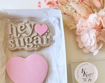 Puede incluir: Una caja de regalo blanca con una tapa transparente contiene tres galletas de azúcar decoradas. Las galletas están decoradas con glaseado rosa y marrón. Las galletas dicen "hey sugar", "I love you" y tienen forma de corazón rosa. La caja tiene una pegatina con el texto "Kelseys Cookies" y las iniciales "KK" en un círculo.