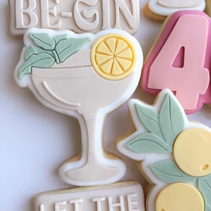 Op de afbeelding: Versierde suikerkoekjes met een cocktailthema. De koekjes bevatten een cocktailglas met een schijfje citroen, de tekst "LET THE 40s BE-GIN" en het nummer "40" in roze.