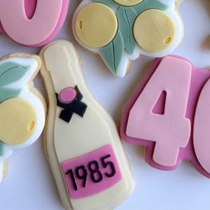 May include: Selection of decorated sugar biscuits with a celebratory theme. Biscuits include champagne bottles with "1985" labels, "LET THE 40s BEGIN" text, and lemon and leaf designs. Pink and white icing.