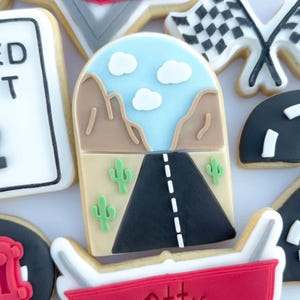 May include: Assortment of decorated sugar cookies with a racing theme. Designs include a road scene with cacti, a checkered flag, and a red emblem with the name "Ottis". The cookies are decorated with white, black, red, blue, and tan icing.