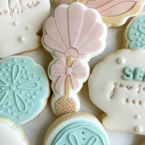 Könnte beinhalten: Auswahl an dekorierten Zuckerkeksen im Babyparty-Thema. Die Kekse umfassen Muschel-, Sanddollar- und Rasselformen, die mit pastellrosa, blauer und weißer Glasur verziert sind. Einige Kekse haben den Text "Baby" und "Sea you soon baby".