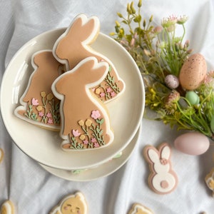 May include: A set of Easter-themed sugar cookies decorated with pastel colours and floral designs. The cookies feature bunnies, chicks, and eggs.