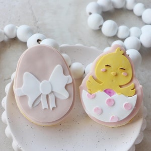 May include: Two decorated sugar cookies on a white plate with a scalloped edge. One cookie is pink with a white bow and the other is white with pink polka dots and a yellow chick inside.