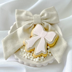 Può includere: Un biscotto marrone chiaro decorato con un fiocco rosa, circondato da codette di perle bianche su un piatto in ceramica marrone chiaro con un fiocco di stoffa beige in cima.