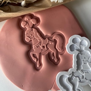 May include: A pink silicone mould with a white plastic mould of a carousel horse. The silicone mould has a raised impression of the horse, while the plastic mould is flat.