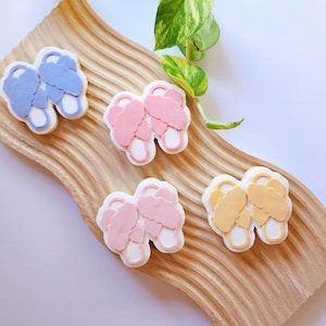 May include: Four pairs of fuzzy slipper shaped charms in pink, light blue, and yellow on a wooden surface.