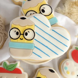May include: A selection of decorated sugar biscuits with a teacher theme. Biscuits include a smiling face, a heart with blue stripes, pencils, apples, and the words "Thank You!" in icing. The biscuits are on a white surface.