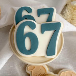 May include: A collection of decorated cookies. One cookie is shaped like the number 67 in teal on a white background. Several cookies are shaped like hands holding a heart. Wooden bead necklaces are in the background.