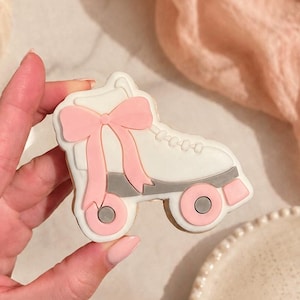 Puede incluir: Una galleta blanca con forma de patín con un lazo rosa y ruedas grises. La galleta está decorada con detalles en rosa y gris. El fondo incluye una tela rosa, un jarrón con flores rosas y un plato.