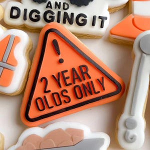Könnte beinhalten: Eine Auswahl an Keksen zum Thema Bau, darunter ein gelber Bagger, Leitkegel und ein Muldenkipper. Einige Kekse tragen die Aufschriften "2 Year Olds Only" und "I'm Digging It". Die Kekse sind mit orangefarbenem, schwarzem, weißem und grauem Zuckerguss verziert.