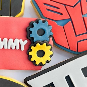 Pode incluir: Variedade de biscoitos de açúcar decorados com designs Transformers. Inclui biscoitos em forma dos símbolos Autobot e Decepticon, as letras "TF" e o número "4". Um biscoito tem o nome "TOMMY".
