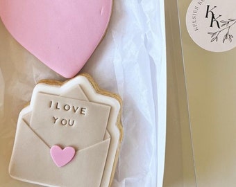 Puede incluir: Una caja de regalo blanca con una tapa transparente contiene tres galletas de azúcar decoradas. Las galletas están decoradas con glaseado rosa y marrón. Las galletas dicen "hey sugar", "I love you" y tienen forma de corazón rosa. La caja tiene una pegatina con el texto "Kelseys Cookies" y las iniciales "KK" en un círculo.