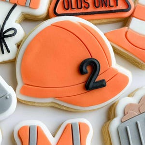 May include: Selection of decorated sugar biscuits with an orange construction theme. Biscuits include a hard hat with the number 2, a safety vest, a road cone, and a sign that reads "2 YEAR OLDS ONLY".