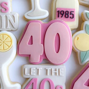 May include: A collection of decorated sugar cookies celebrating a 40th birthday. The cookies feature a champagne bottle, cocktail glasses, and the number 40 in pink and white. Some cookies have the text "LET THE 40s BE-GIN".