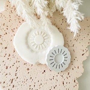 Puede incluir: Dos moldes de cerámica blanca en forma de sol con un diseño en relieve. El molde más grande tiene un centro sólido, mientras que el molde más pequeño tiene un centro hueco. Los moldes están sobre una superficie texturizada rosa.