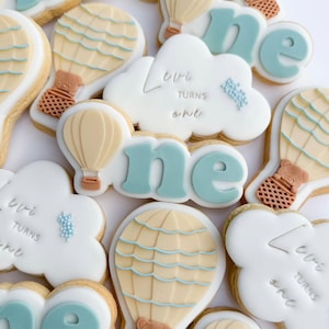 Puede incluir: Un conjunto de galletas de azúcar decoradas con un tema de globo aerostático azul y blanco. Las galletas están decoradas con las palabras "Levi cumple un año" y "uno".