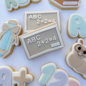 Può includere: Assortimento di biscotti di zucchero decorati a tema scolastico. I biscotti includono le lettere "ABC", numeri, una matita con un fiocco, una pila di libri, un verme con gli occhiali e una lavagna con scritto "ABC 2+2=4".