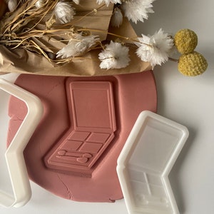 Può includere: Due stampini per biscotti in plastica bianca a forma di palette di ombretti. Uno stampino è chiuso e l'altro è aperto, mostrando i singoli scomparti per ombretti.